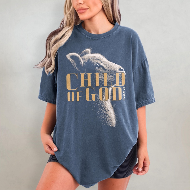 May include: A navy blue t-shirt featuring a detailed illustration of a calf's head with the text "CHILD OF GOD" in gold lettering. The shirt also includes the text "PSALM 23". The t-shirt has short sleeves and a relaxed fit.