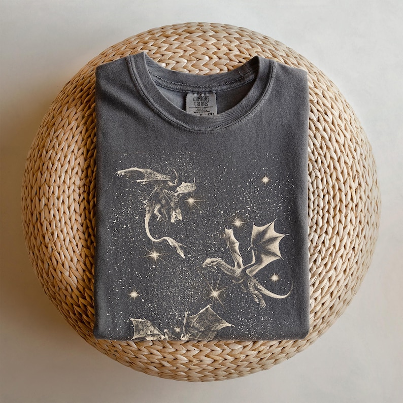 May include: A dark gray t-shirt with a graphic print of two dragons flying through a starry sky. The shirt has a crew neck and short sleeves. The text "COMFORT COLORS" is printed on the tag.