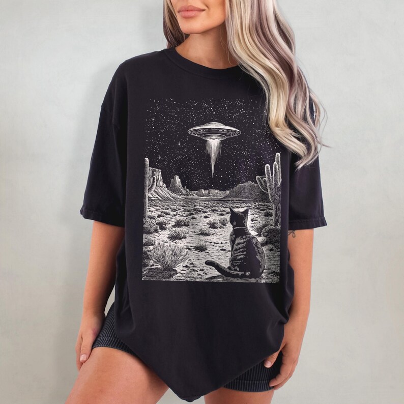 May include: Black oversized t-shirt with a graphic print of a cat sitting in a desert landscape looking up at a UFO in the night sky.
