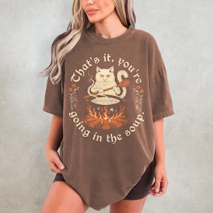 May include: Brown t-shirt with a cat graphic stirring soup over a fire, surrounded by text that reads "That's it, you're going in the soup."
