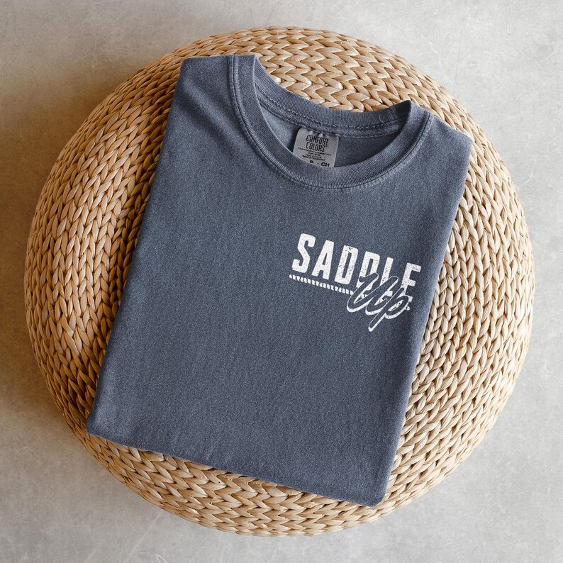 May include: A slate blue t-shirt with the words "SADDLE UP" printed in white. The shirt is folded and resting on a round, woven, light brown surface. The shirt has a crew neck and short sleeves.