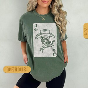 Puede incluir: Una camiseta verde con un gr&aacute;fico blanco de un esqueleto con un sombrero de vaquero y un pa&ntilde;uelo. El gr&aacute;fico es una carta de juego con la letra 'J' y un s&iacute;mbolo de diamante en la esquina superior izquierda. Las palabras 'COMFORT COLORS' y 'MOSS' son visibles en la imagen.