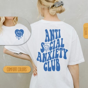 K&ouml;nnte beinhalten: Wei&szlig;es T-Shirt mit blauem Text, der "Anti Social Anxiety Club" in einer Retro-Schriftart lautet. Ein blaues, tropfendes Smiley-Gesichtsherz befindet sich im Text. Das Shirt ist in Wei&szlig; und in Komfortfarben erh&auml;ltlich.