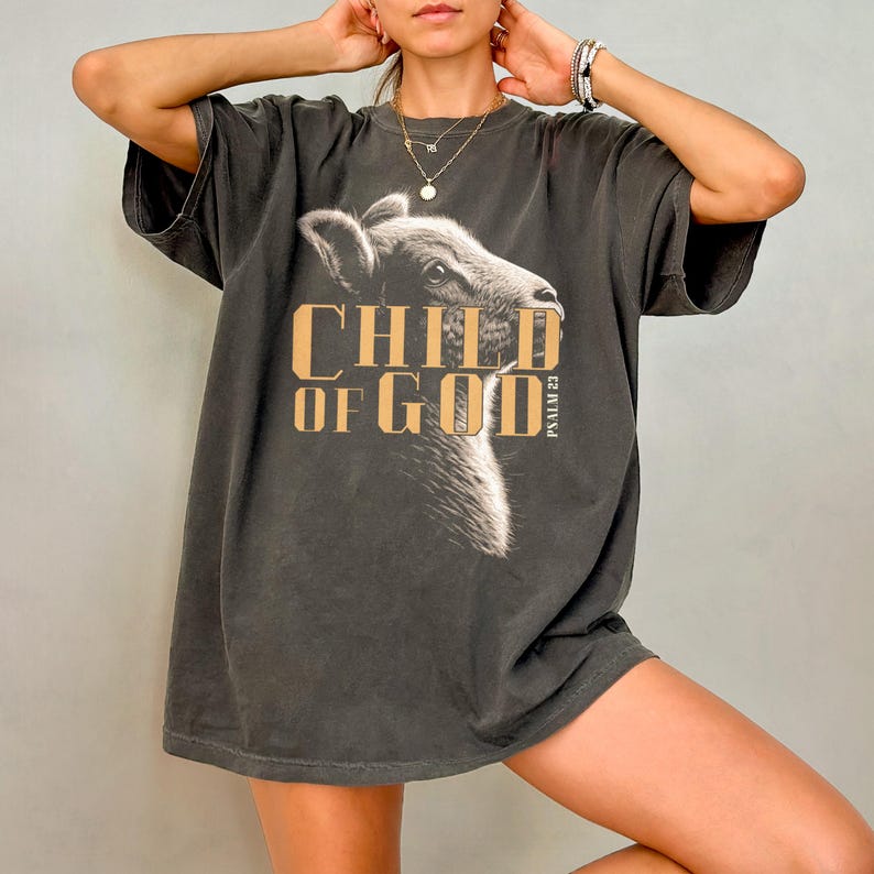 May include: Dark gray t-shirt featuring a graphic of a calf's head with the text "CHILD OF GOD" in a gold color. The shirt also includes the text "PSALM 23". The shirt has short sleeves and a relaxed fit.