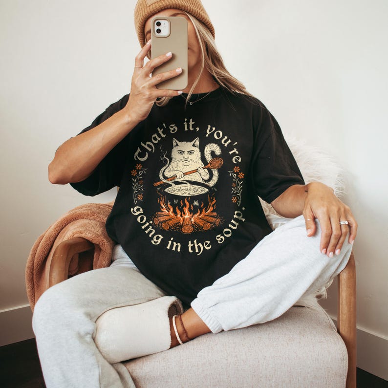 May include: Black t-shirt with a humorous graphic of a cat stirring a pot over a fire, with the text "That's it, you're going in the soup." The design is surrounded by floral accents.