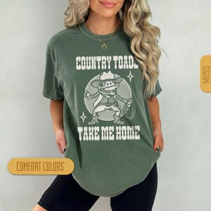 May include: A green t-shirt with a white graphic of a cartoon frog wearing a cowboy hat and holding a lasso. The text "COUNTRY TOAD TAKE ME HOME" is printed on the shirt.
