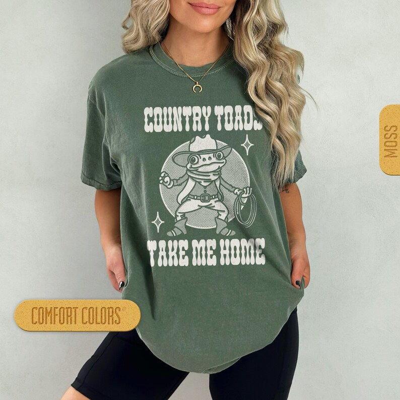 May include: A green t-shirt with a white graphic of a cartoon frog wearing a cowboy hat and holding a lasso. The text "COUNTRY TOAD TAKE ME HOME" is printed on the shirt.