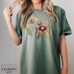 May include: A green t-shirt with a floral print featuring a variety of flowers in shades of red, orange, yellow, and green. The flowers are arranged in a bouquet-like shape and are set against a light green background.