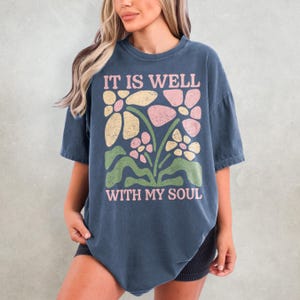 May include: A navy blue t-shirt with the text "IT IS WELL WITH MY SOUL" in pink. The shirt features a floral design in pink, yellow, and green. The t-shirt is a relaxed fit.