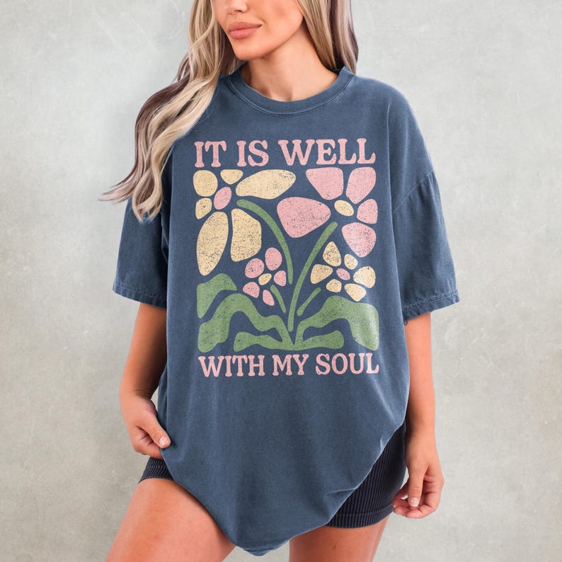 May include: A navy blue t-shirt with the text "IT IS WELL WITH MY SOUL" in pink. The shirt features a floral design in pink, yellow, and green. The t-shirt is a relaxed fit.