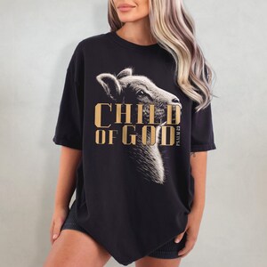 May include: Black t-shirt featuring a detailed illustration of a lamb's head with the text "CHILD OF GOD" in gold lettering. The words "PSALM 23" are printed vertically on the right side of the shirt. The shirt has short sleeves.