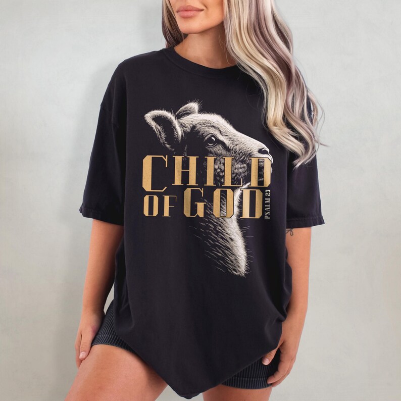 May include: Black t-shirt featuring a detailed illustration of a lamb's head with the text "CHILD OF GOD" in gold lettering. The words "PSALM 23" are printed vertically on the right side of the shirt. The shirt has short sleeves.