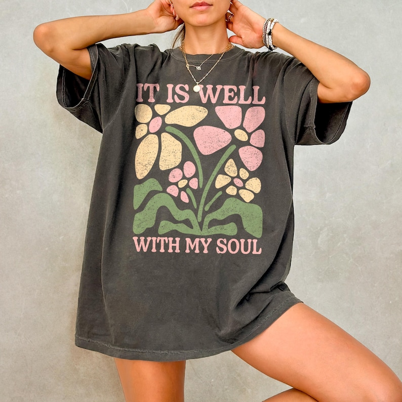 May include: Dark gray t-shirt with the text "IT IS WELL WITH MY SOUL" in pink and white. The shirt features a floral design in pink, yellow, and green. The shirt is a relaxed fit.