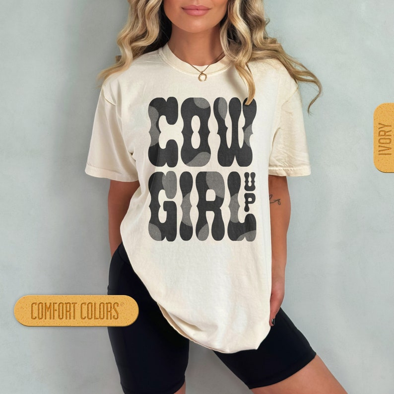 May include: A white t-shirt featuring a black graphic that reads "COWGIRL" in a retro font. The graphic is made up of black and grey cow spots. The shirt is being modelled by a person with long brown hair. The shirt is available in ivory and comfort colours.