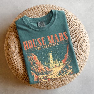 May include: A teal t-shirt with the text "HOUSE MARS THE INSTITUTE" in orange lettering. The graphic depicts a castle scene with red rock formations and a starry sky. The shirt is folded on a woven, circular mat.