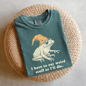 May include: Teal t-shirt featuring a whimsical illustration of a frog wearing a wizard hat. The frog is cream-colored with detailed shading. The orange hat has star accents. The text below the frog reads, "I have to say weird stuff or I'll die."