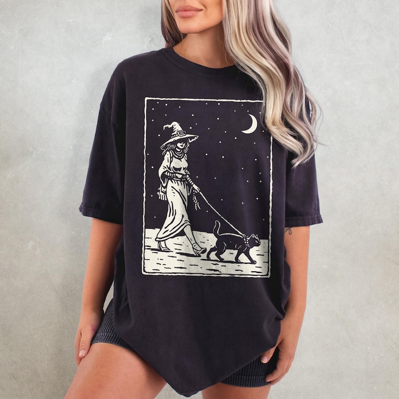 May include: Dark navy blue t-shirt featuring a black and white graphic of a witch walking a black cat under a starry night sky. The witch is wearing a hat and long dress. The design is framed in a white rectangle.