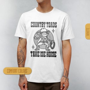 May include: White t-shirt with a black and white illustration of a frog wearing a cowboy hat and holding a lasso. The text "COUNTRY TOADS TAKE ME HOME" is printed on the shirt.