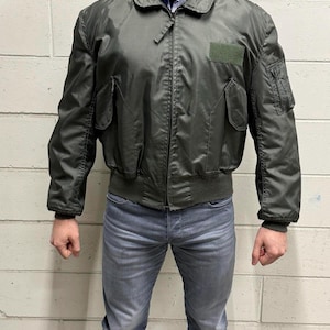 Puede incluir: Una chaqueta bomber verde oscuro con cierre de cremallera y un parche en la manga izquierda. La chaqueta tiene un cuello y puños de canalé.