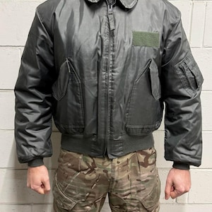 Puede incluir: Chaqueta bomber gris oscuro con cremallera, cuello y múltiples bolsillos. La chaqueta tiene un parche verde en el pecho y puños y dobladillo acanalados. La persona lleva pantalones de camuflaje.