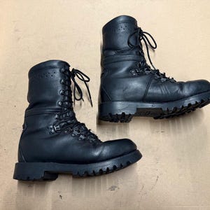 Könnte beinhalten: Paar schwarze Leder-Kampfstiefel mit dicken, profilierten Sohlen und Schnürverschlüssen. Die Stiefel haben mehrere Ösen und Metallbeschläge. Die Stiefel stehen auf einer hellbraunen Oberfläche.