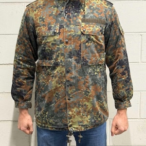 May include: A camouflage jacket with a hood, in a pattern of green, brown, and grey spots. The jacket has multiple pockets, button fastenings, and a drawstring at the waist. The sleeves have buttoned cuffs.