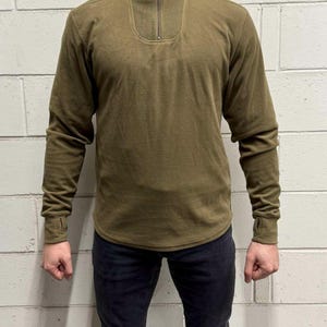 Könnte beinhalten: Olivgrüner Fleece-Pullover mit hohem Kragen und Viertel-Reißverschluss. Das langärmlige Shirt hat eine lockere Passform und gerippte Bündchen. Das Model trägt eine schwarze Jeans. Hintergrund: weiße Backsteinmauer.