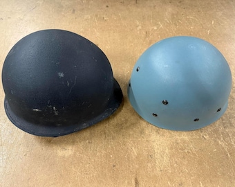 May include: Two military-style helmets. One is black, the other is a light blue-gray. Both have a rounded shape with a brim and rivets. The helmets are likely for protection or historical reenactment.