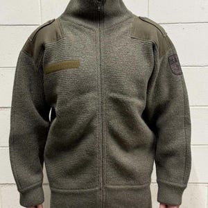 May include: Olive green zip-up sweater with a high collar. The sweater features shoulder patches, a small rectangular patch on the chest, and a patch on the right sleeve. The sweater has a textured knit design.