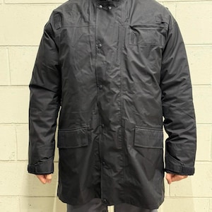 May include: A black, long coat with a high collar, button and zipper closure, and multiple pockets. The coat appears to be made of a water-resistant material. The sleeves have adjustable cuffs. The coat is worn over gray shorts.