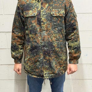 May include: A camouflage jacket in shades of green, brown, and black. The jacket has multiple pockets, a hood, and a drawstring at the waist. The person is wearing blue jeans.