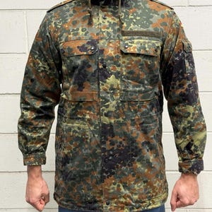 Op de afbeelding: Een camouflagejas in tinten groen, bruin en zwart. De jas heeft een capuchon, meerdere zakken en een trekkoord in de taille. De jas is dichtgeknoopt en heeft een geribbelde kraag.