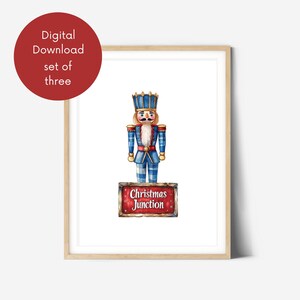 Könnte beinhalten: Ein gerahmter Druck mit einer Aquarellillustration eines traditionellen blau-roten Nussknackersoldaten. Der Nussknacker steht auf einem Schild mit der Aufschrift "Christmas Junction". Ein roter Kreis oben links zeigt "Digitaler Download, Set von drei".