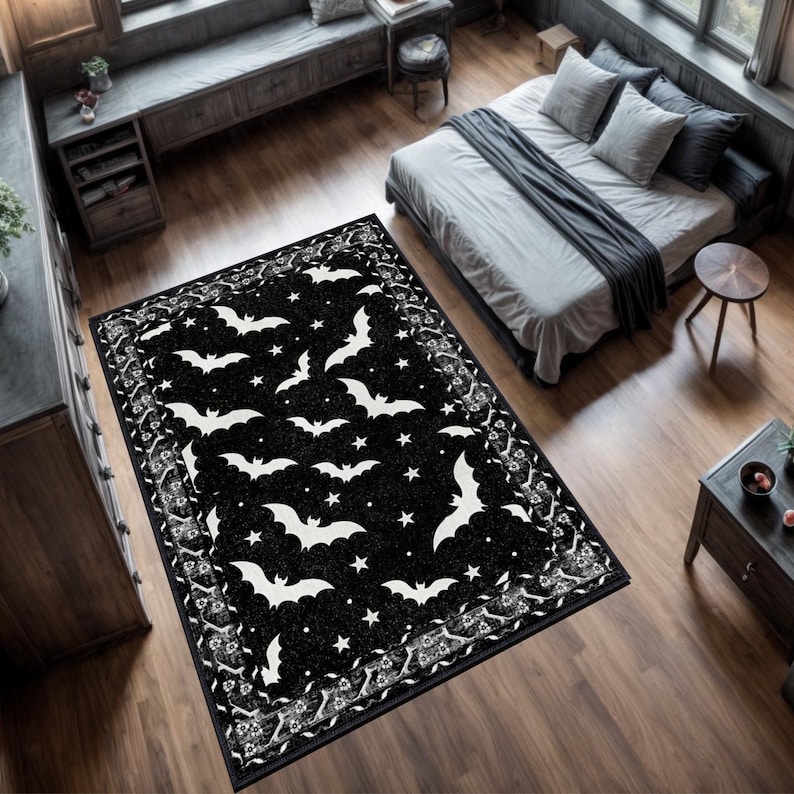 May include: A black rectangular rug featuring a pattern of white bats and stars. The rug has a decorative border with floral and scroll designs. The rug is placed on a wooden floor in a bedroom setting.