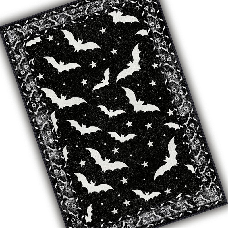 May include: Black rectangular rug with a Halloween theme. The rug features white bats and stars on a black background. The border has a white floral pattern on a black background.