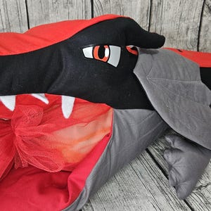 Könnte beinhalten: Ein Plüschdrachenspielzeug in Rot, Schwarz und Grau. Der Drache hat einen roten Kopf mit schwarzen Akzenten, weißen Zähnen und großen Augen. Graue Flügel und Füße erstrecken sich vom Körper. Ein roter und orangefarbener Tüllrock ist sichtbar.