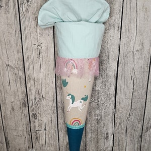 Könnte beinhalten: Ein hellblauer und weißer Einhorn-Schulkegel mit rosa-weißer Spitzenborte. Der Kegel hat ein Regenbogen-Design und ein Einhorn mit einem Herz auf der Seite. Der Kegel steht auf einer weißen, flauschigen Oberfläche.