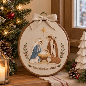 Puede incluir: Un bastidor de bordado circular con una escena de natividad que muestra a María, José y el niño Jesús en un pesebre. El diseño incluye una estrella, un cordero y una vaca, con el texto EMMANUEL'S ANNA bordado abajo y un lazo de tela beige.