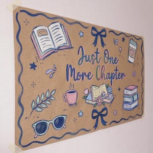 May include: A rectangular tan poster with a dark blue wavy border featuring the text Just One More Chapter in blue. Illustrations include an open book, a stack of books, a coffee cup, a drink tumbler with a heart, sunglasses, and blue bows.