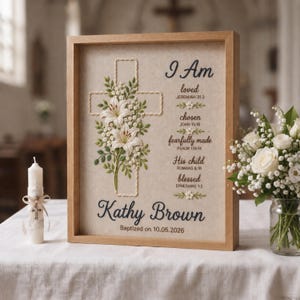 Peut inclure: Une broderie encadrée sur tissu beige représentant une croix blanche ornée de lys brodés et de feuilles vertes. Le texte indique I Am loved, chosen, fearfully made, His child, blessed, Kathy Brown, et Baptized on 10.05.2026.