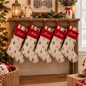 Peut inclure: Cinq chaussettes de Noël beiges avec un motif répétitif de sapins verts et de nœuds rouges. Chaque chaussette possède un revers rouge brodé avec les prénoms Dad, Mom, Lucas, Jenny et Laura, contenant des cadeaux et des sucres d'orge.