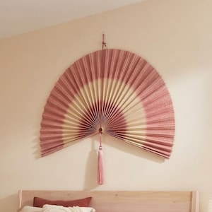 Peut inclure: Un grand éventail mural semi-circulaire présentant un design plissé avec des bandes alternées de couleurs rose poudré et crème. L'éventail est suspendu par un cordon fin et comprend un pompon rose poudré assorti suspendu au centre.