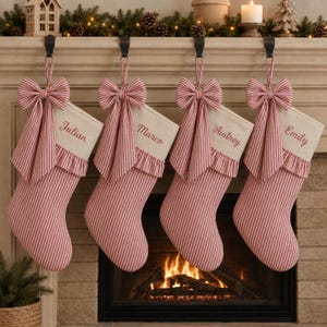 May include: Four Christmas stockings made of cream-coloured fabric with red vertical stripes. Each stocking features a ruffled cuff, a large matching red and white striped bow, and the embroidered names Julian, Marco, Audrey, and Emily in red.