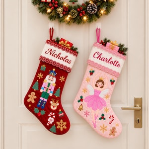 Könnte beinhalten: Zwei personalisierte Weihnachtsstrümpfe hängen an einer Tür. Der linke Strumpf ist rot mit Nussknacker-, Lebkuchenmann- und Schneeflockenstickerei. Der rechte Strumpf ist rosa mit Ballerina-, Stechpalmen- und Zuckerstangenstickerei. Beide haben weiße Bündchen.