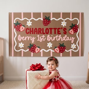 Puede incluir: Una pancarta marrón con rayas rosas y un borde blanco ondulado, con fresas rojas y flores blancas. La pancarta dice "CHARLOTTE'S berry 1st birthday" en letras rojas y rosas. Una niña con un vestido con temática de fresas está frente a un regalo.