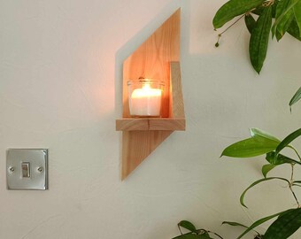 May include: A wooden candle holder with a geometric design, featuring a shelf to hold a candle. The candle holder is mounted on a white wall, and a lit candle is placed on the shelf.