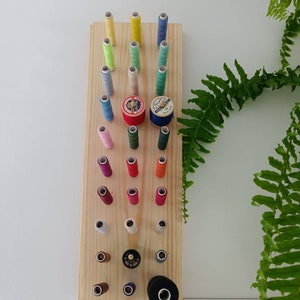 May include: A wooden thread rack with a variety of colored thread spools. The rack has 25 slots for spools and is made of light wood. The spools are arranged in rows and columns.
