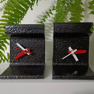 May include: Two black metal bookends shaped like a railway track with a clock face on each. The clock hands are red and white.