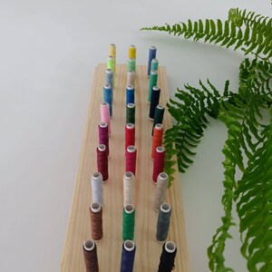 May include: A wooden spool holder with 25 spools of thread in various colors, including red, green, blue, and brown. The spools are arranged in rows on the holder.