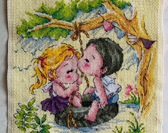 May include: Cross-stitch artwork depicting a couple kissing under a tree. The artwork features a yellow background, with the couple in the center, and a tree with green leaves and a brown trunk above them. The couple is sitting on a dark green surface.